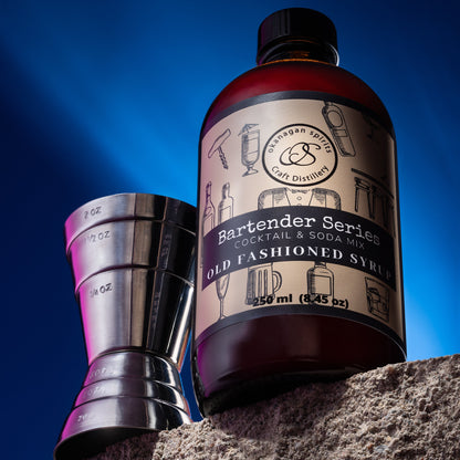 Bottle of Old Fashioned Syrup with a cocktail shaker on a stone ledge against a blue sky.