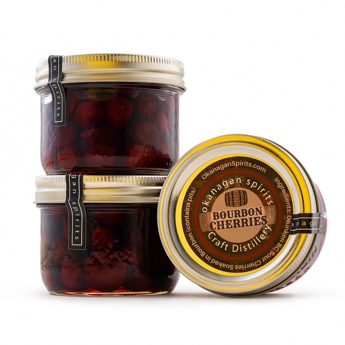 Two glass jars of dark red preserved cherries with gold lids, alongside one open jar showing the round label. The label reads Okanagan Spirits Bourbon Cherries Craft Distilled.