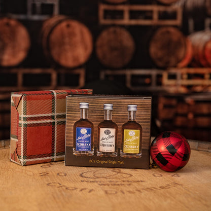 Three miniature bottles of whisky in a decorative box with plaid design, surrounded by wooden barrels.